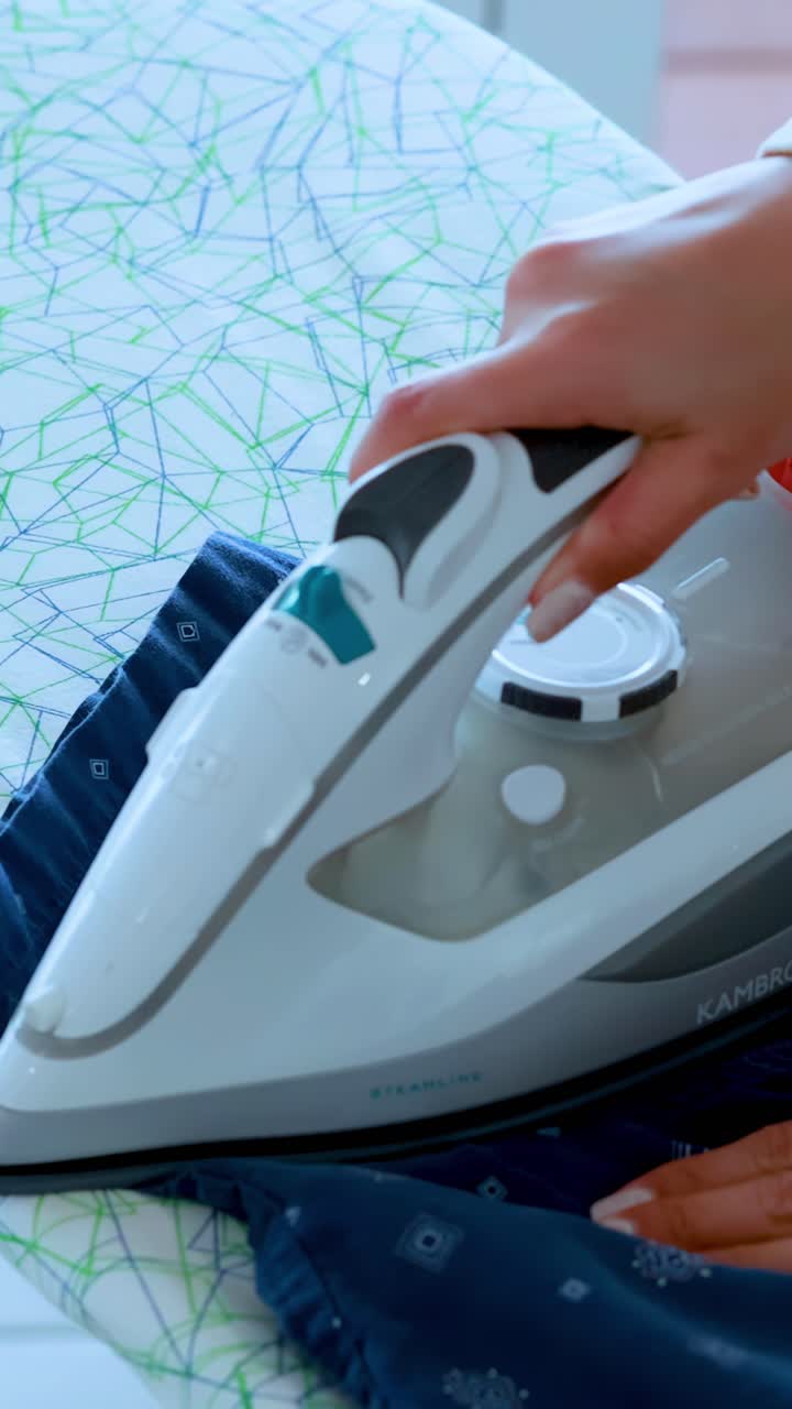 A woman smooths and irons a blue patterned shirt on a modern ironing board in bright, natural light, using careful, precise movements