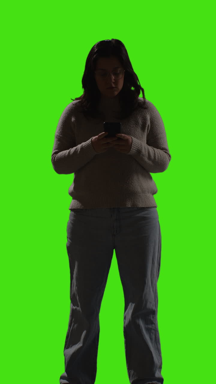 disparo de video vertical de una mujer joven usando un teléfono móvil para enviar mensajes de texto con una iluminación de baja clave de pie contra la pantalla verde