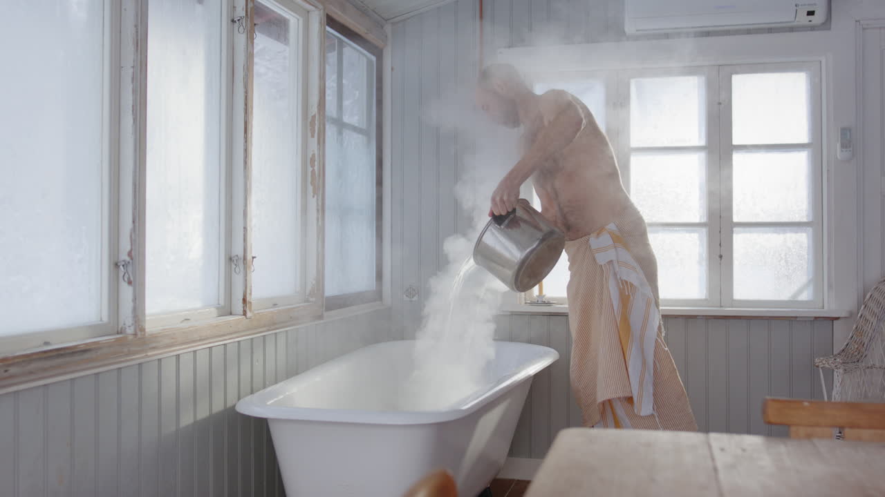 Bare chested man in towel pours boiling hot water from woodstove into steam bath