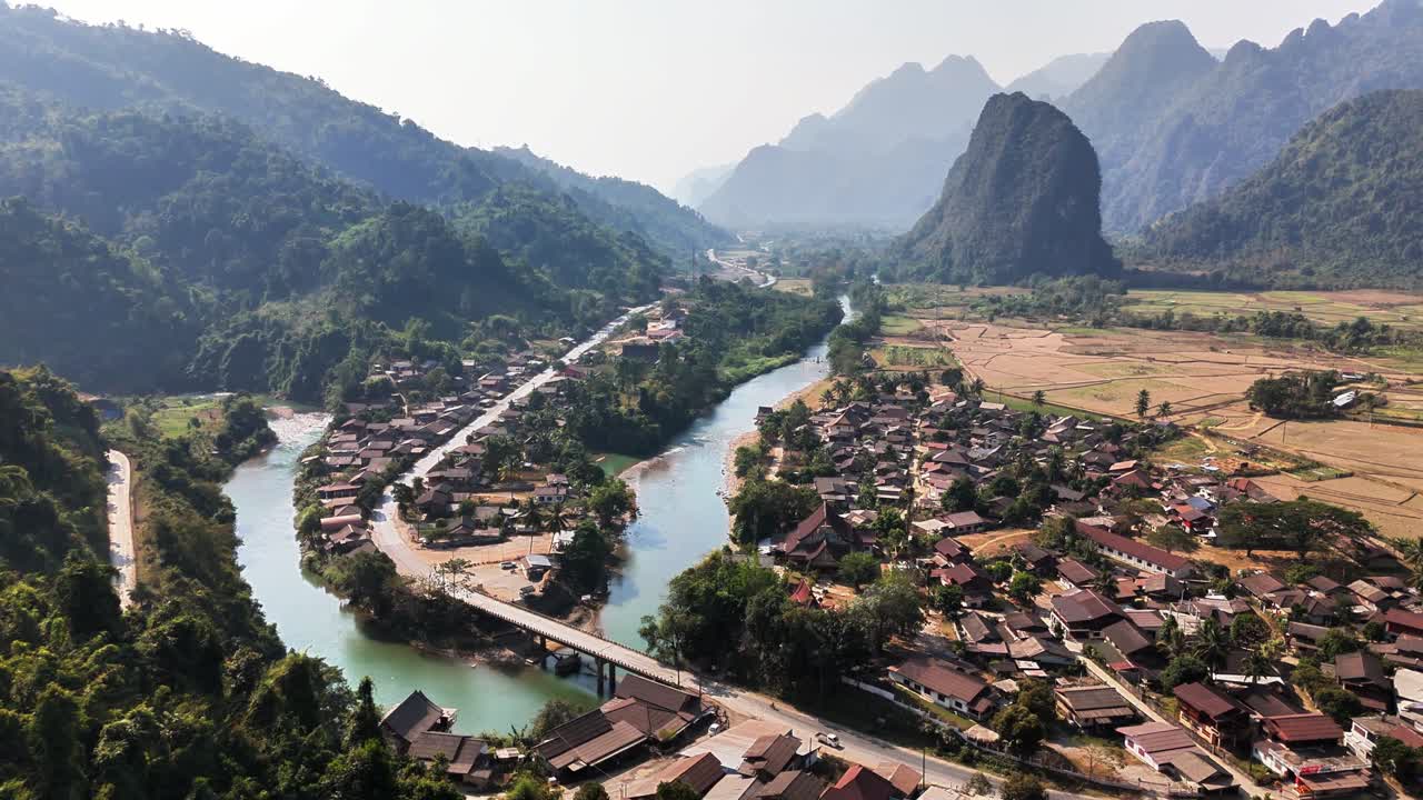 Tranquillo villaggio rurale di Pha Tang annidato lungo un fiume serpeggiante tra montagne lussureggianti e terreni agricoli a Vang Vieng, Laos. La vista dall'alto evidenzia ponti, case tradizionali e paesaggi naturali