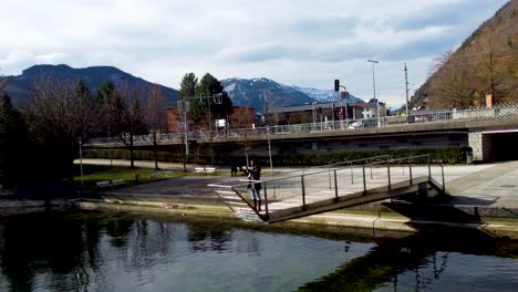 Tourist-taking-photo-on-a-footbridge-in-Ebensee,-Traunsee-lake-nearby,-serene-mountain-backdrop,-sunny-day