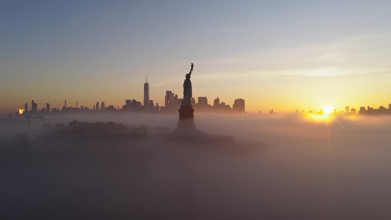 Luftoptagelse, der daler ned over Frihedsgudinden dækket af morgentåge med silhuetten af Manhattans skyline i baggrunden ved solopgang, New York.