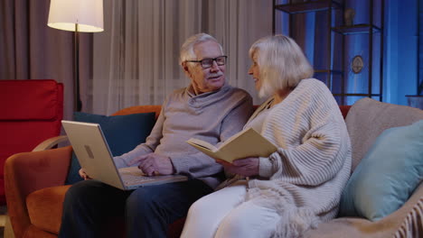 Senior-grandparents-couple-reading-book,-using-laptop-pc-on-couch-in-night-living-room-at-home