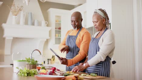 pareja de ancianos afroamericanos en delantales preparando una comida usando una tableta en la cocina, cámara lenta