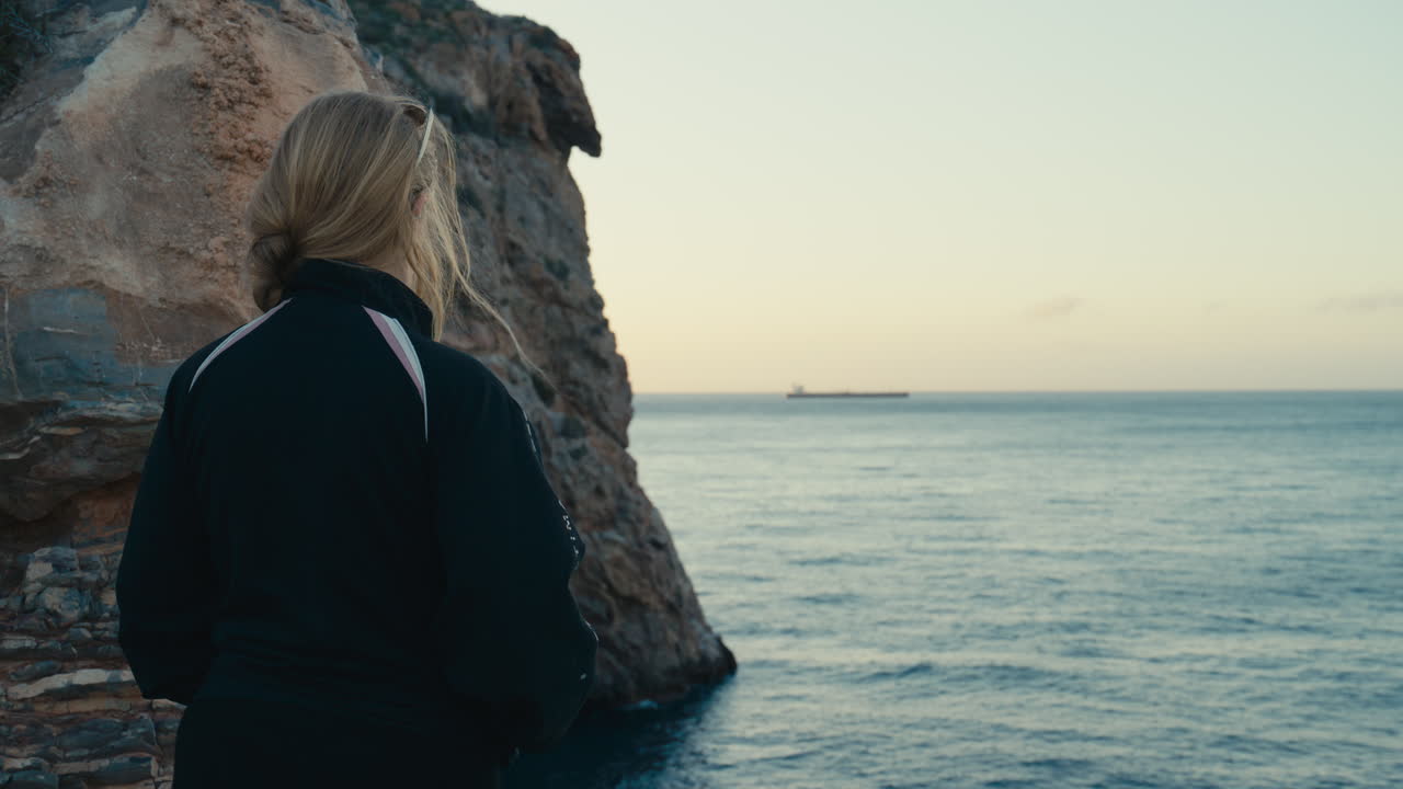 A woman stands by a rocky cliff, gazing out over the calm ocean during golden hour. Peaceful and reflective atmosphere with a sense of solitude and freedom