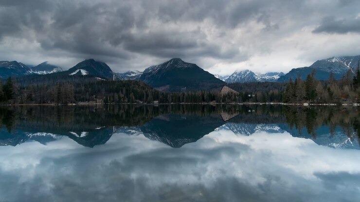 Ampia inquadratura 4K dei monti Tatra innevati, del lago calmo di Štrbské Pleso, di una pineta, di un cielo cupo e di alberi illuminati dal sole—perfetto per riprese di viaggio, natura, resort, meditazione e paesaggi
