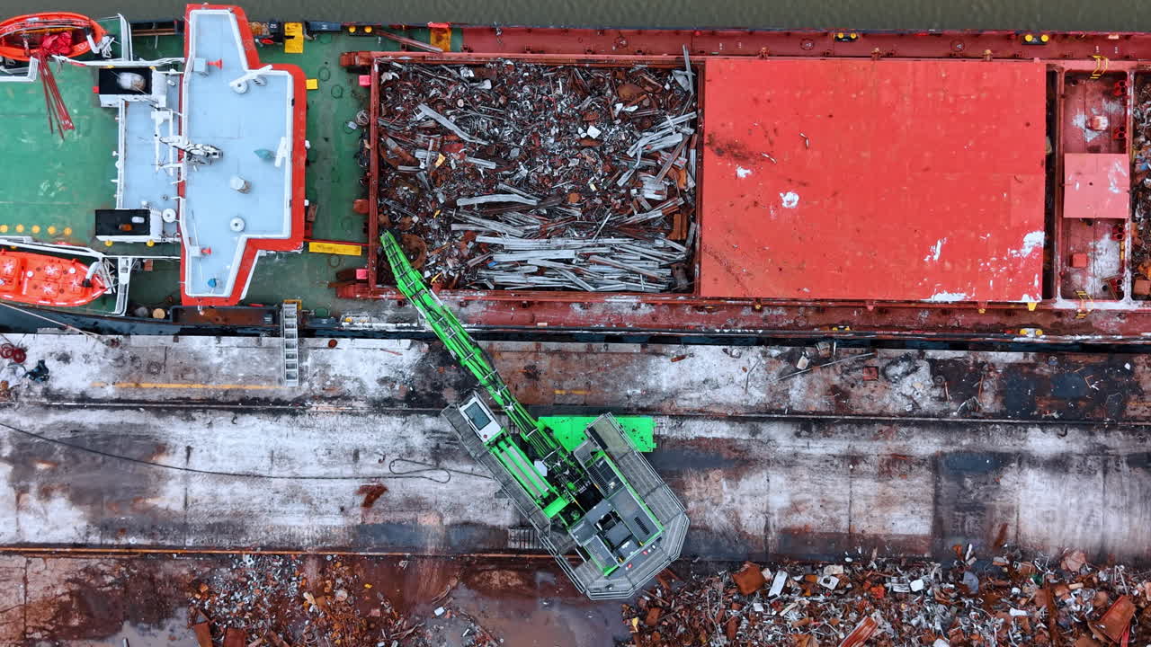 Vista dall'alto di una gru verde che carica rottami metallici su una nave da carico. Manipolatore gru industriale verde che sposta rottami metallici nella stiva di una nave da carico rossa presso il molo del cantiere navale