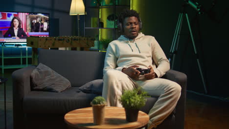 african american player relaxing at home sitting down on couch, excited to play games and enjoy leisure time. person on sofa in a decorated living room, playing video games on console. camera b.