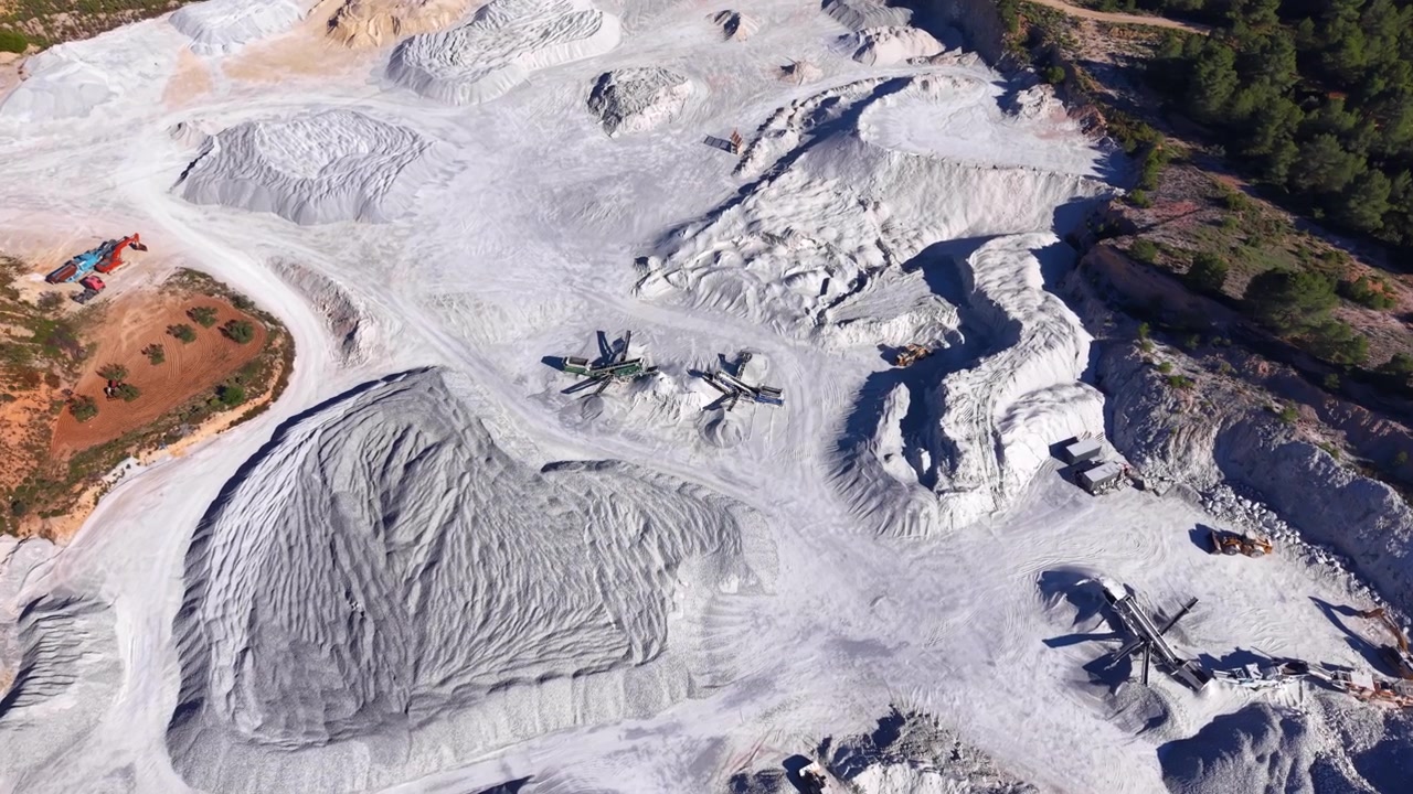 Vista aérea de una gran cantera de yeso con depósitos minerales blancos, maquinaria pesada, excavadoras y caminos de tierra sinuosos, rodeada de terreno rocoso y pinos.