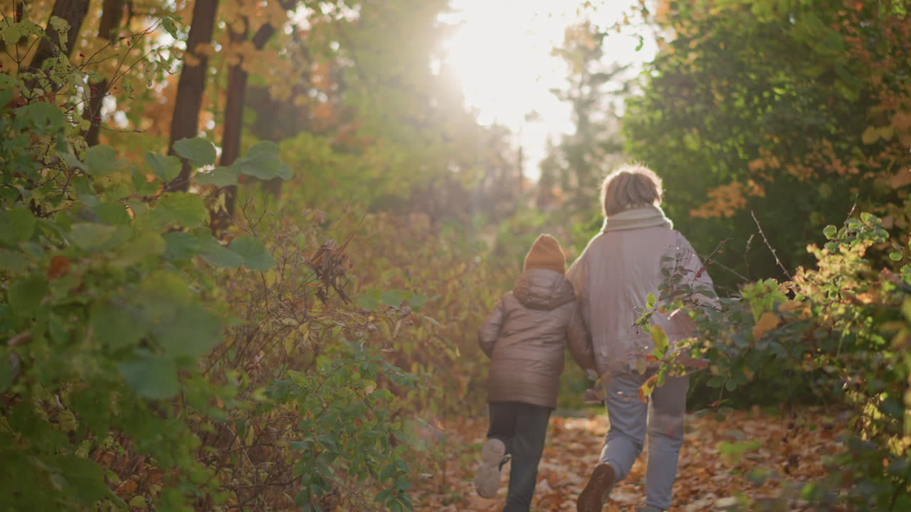 back view of adult and child dressed in puffy jackets running together through autumn forest trail, warm sunlight filtering through colourful trees, capturing bond and seasonal joy in natural setting