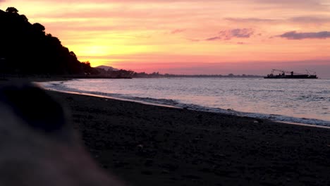 watching stunning pink orange sunset with ocean waves rolling in, traffic driving around the coastline and a large ship dredging boat on tropical island timor leste, southeast asia