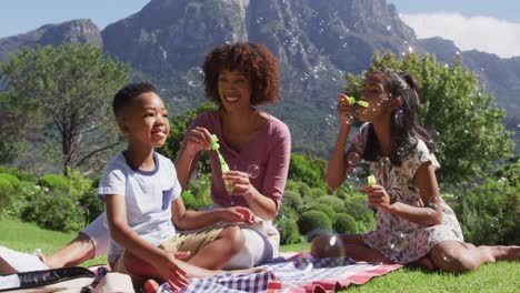 feliz madre afroamericana, hija e hijo sentados al aire libre en una manta, soplando burbujas