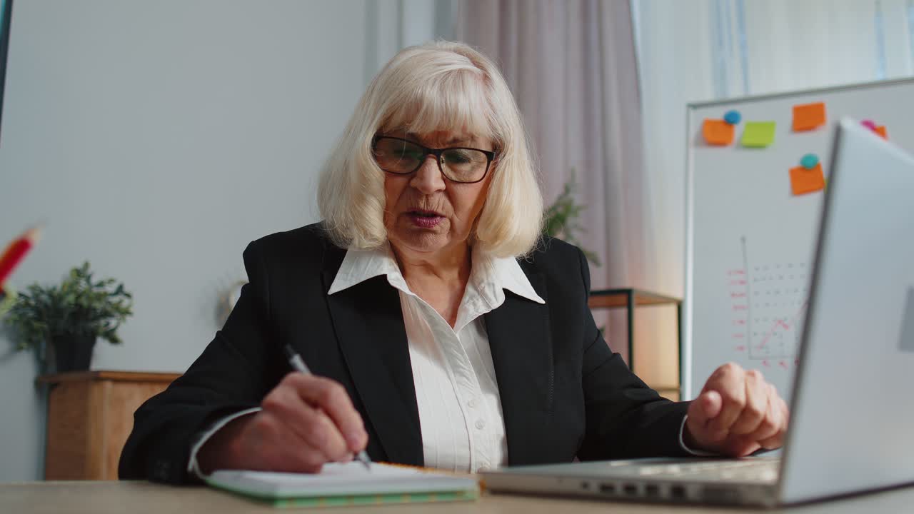 Senior elderly business office woman elearning online writing notes from laptop live webinar online