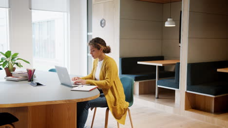 Mujer-Adulta-Joven-Que-Trabaja-En-Una-Computadora-Portátil-Sentada-En-Una-Oficina-Mientras-Su-Colega-Entra-A-La-Habitación,-Con-La-Mano