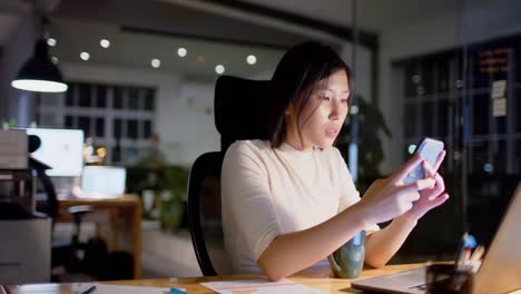 Fröhliche-Asiatische-Geschäftsfrau-Am-Schreibtisch-Mit-Smartphone-Im-Büro-Bei-Nacht,-Zeitlupe