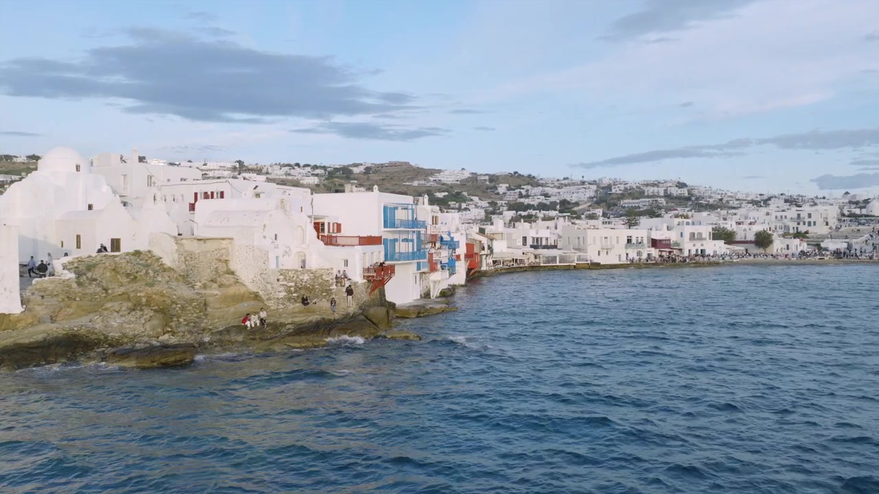Edifici imbiancati a calce di Mykonos arroccati su un promontorio roccioso sopra il mare, con la costa della città e case collinari che si estendono in lontananza.