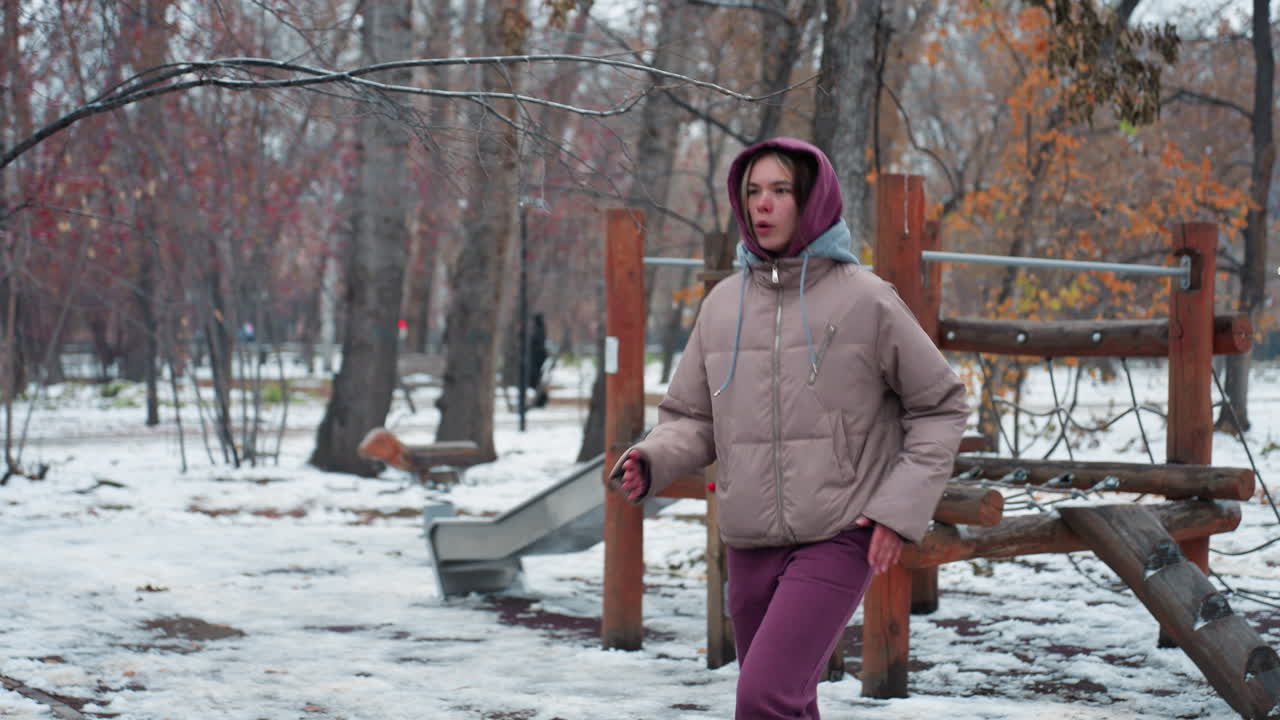 Girl in winter outfit warms up in cold outdoor park in urban setting, walking near playground equipment surrounded by snow and leafless trees, dressed in hoodie, jacket and boots