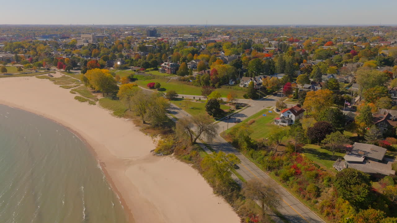 Drone aerial flying over Lake Michigan and sandy beach, pushing toward charming Sheboygan, Wisconsin neighborhood houses surrounded by colorful autumn trees at peak fall color