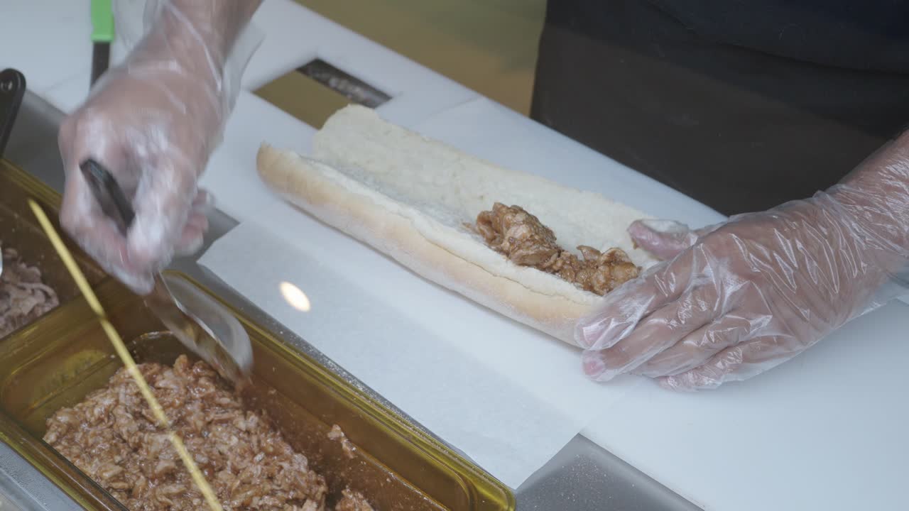Food worker wearing gloves uses a spoon to fill a sandwich roll with cooked, saucy meat over a white prep counter.