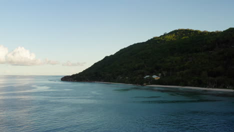 Aerial-shot-of-the-coastline-of-a-lush,-beautiful-green-mountainous-island-surrounded-by-calm-ocean-waters-and-clear-blue-skies