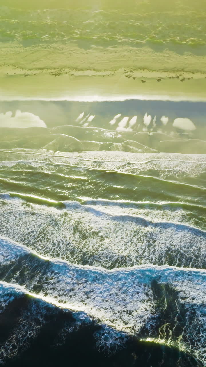 White foamy little waves splashing on the sandy beach at daytime. Bright sun lighting the coast of the ocean. Top view. Vertical video