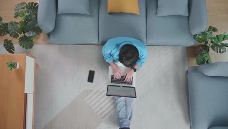 top view of a man typing on a laptop while sitting on the floor with smartphone in front of a sofa at home