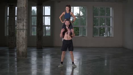 Athletic-shirtless-man-crouches-with-a-girl-on-his-shoulders-in-empty-loft-studio.-Does-squats-with-his-partner-on-shoulders.-Slow-motion.-Front-view