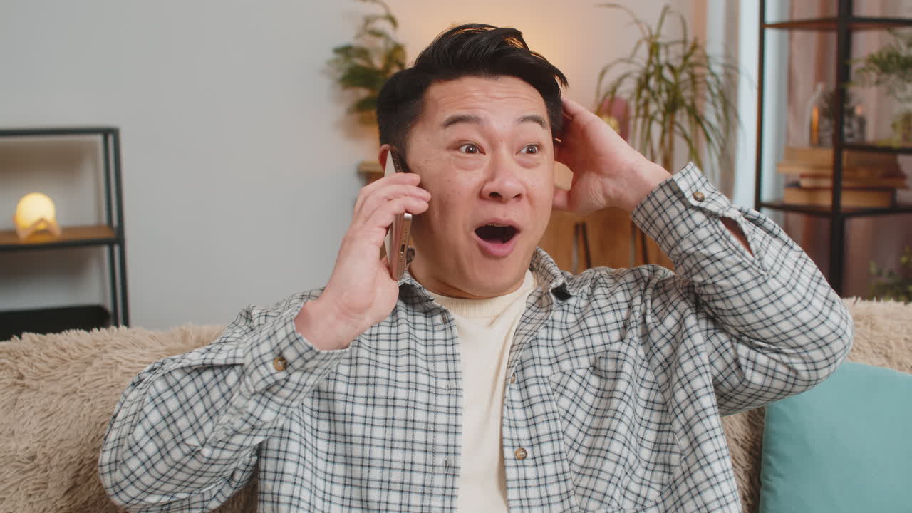 Smiling young chinese man making fun phone conversation with friends sitting on couch in living room