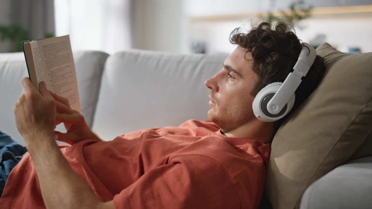Headphones man reading book chilling sofa at home closeup. Guy laying couch