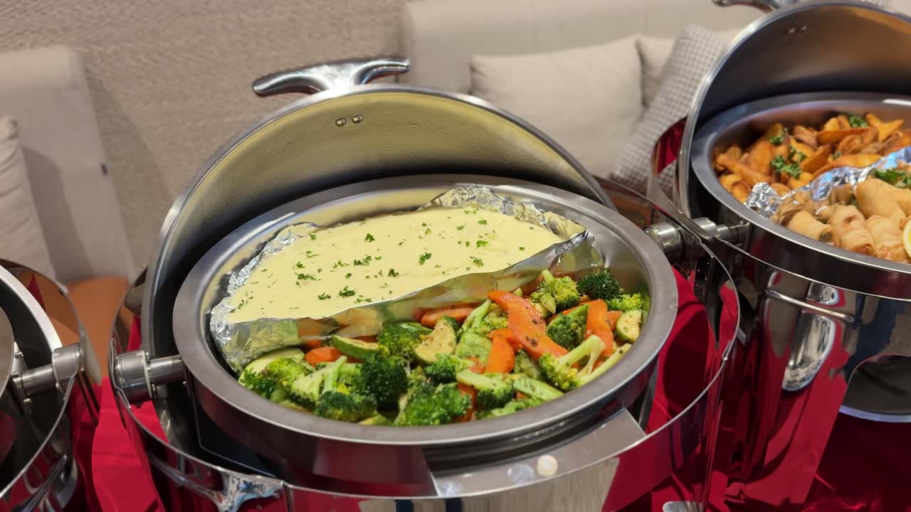 Primo piano di scaldavivande a buffet con pesce al forno guarnito con erbe aromatiche, broccoli e carote al vapore e antipasti fritti con patatine, esposti su un tavolo da catering all'interno.