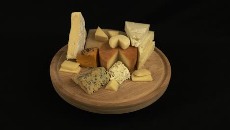 Wood-cutting-board-with-assorted-cheese-rotates-on-black-background