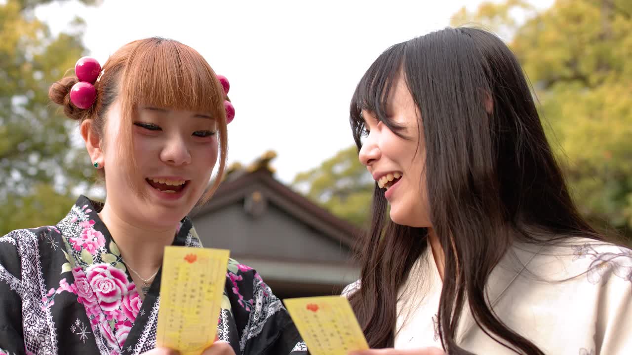 日本の神社で、伝統的な着物を着て微笑む二人の女性が黄色いおみくじを読む