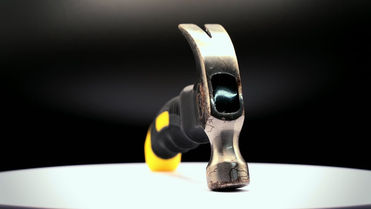 Close-up static shot of a small rusty claw hammer with black and yellow rubber handle rotating 360 degrees on a platform under studio lighting