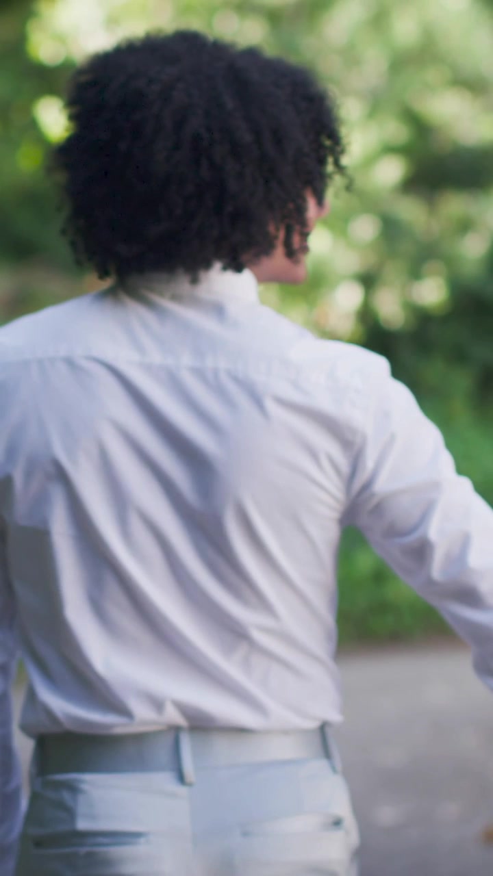 Vertical video: Walking man in white shirt turning to nearby partner on leafy path, holding hands