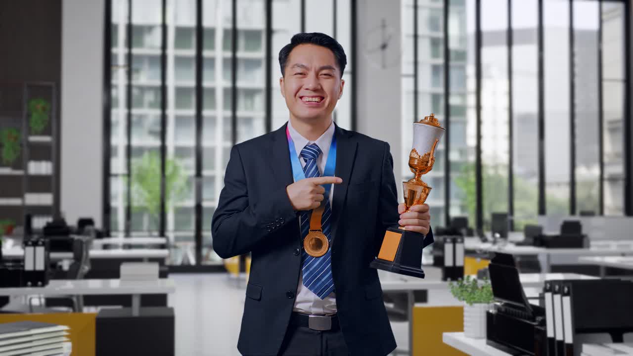 Asian Business Man In A Suit And Tie With A Gold Medal Pointing To A Gold Trophy In His Hand Being Proud Winning As The First Winner In Office
