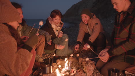 grupo de amigos adolescentes sentados ao redor da fogueira enquanto assam marshmallows e conversam à beira-mar