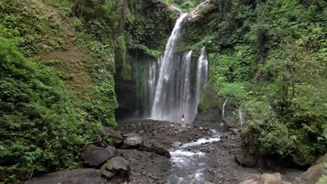 disparo aéreo hacia adelante entre colinas cubiertas de vegetación y una niña frente a una cascada, lombok