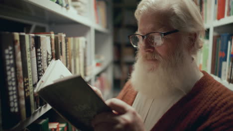 senior bearded man in glasses choosing book to read from library shelf