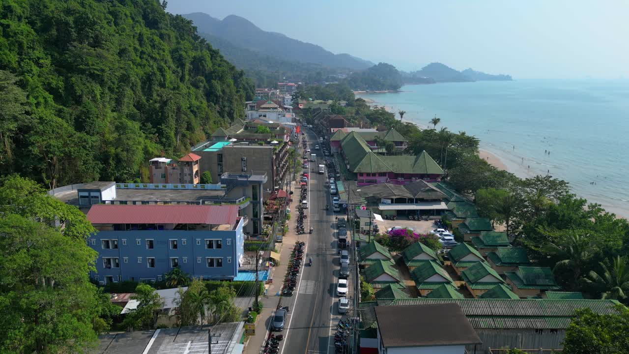 Cars and motorbikes driving on a busy street in Koh Chang, Thailand, with hotels, shops, and the beach in the background. Best aerial view flight tilt down drone