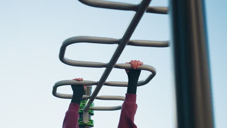fair skinned woman wearing gloves grips silver monkey bars overhead under clear sky in urban park workout showcasing strength focus determination during outdoor calisthenics session