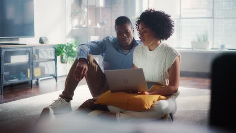 black couple use laptop computer, while sitting on living room floor in their stylish apartment. girlfriend and boyfriend talk, do online shopping on internet, choose product to order online