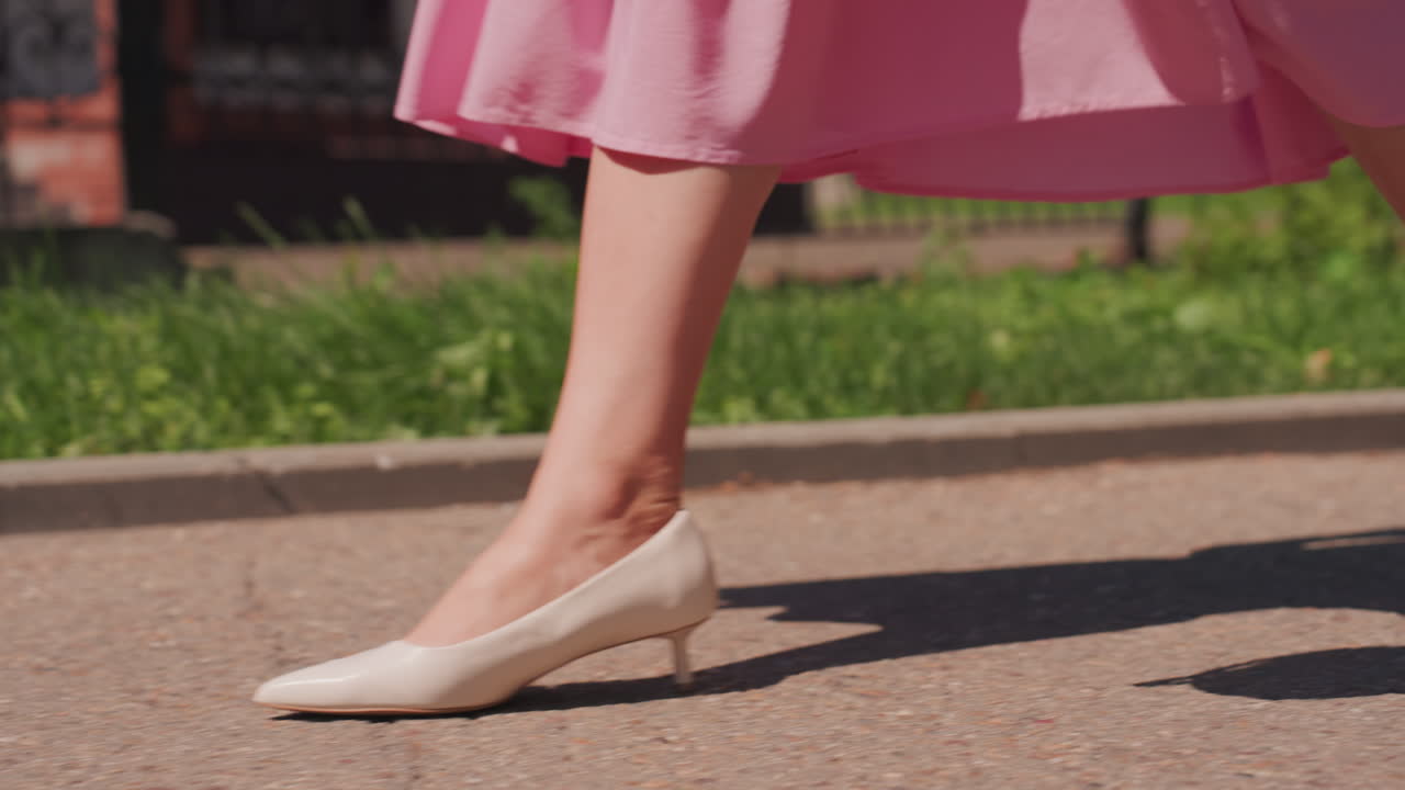 Primo piano delle gambe di una donna che cammina su un marciapiede illuminato dal sole, gonna rosa che ondeggia e décolleté bianche che si muovono delicatamente, leggera ombra sull'asfalto, verde del parco sullo sfondo, andatura leggiadra e rilassata con borsa