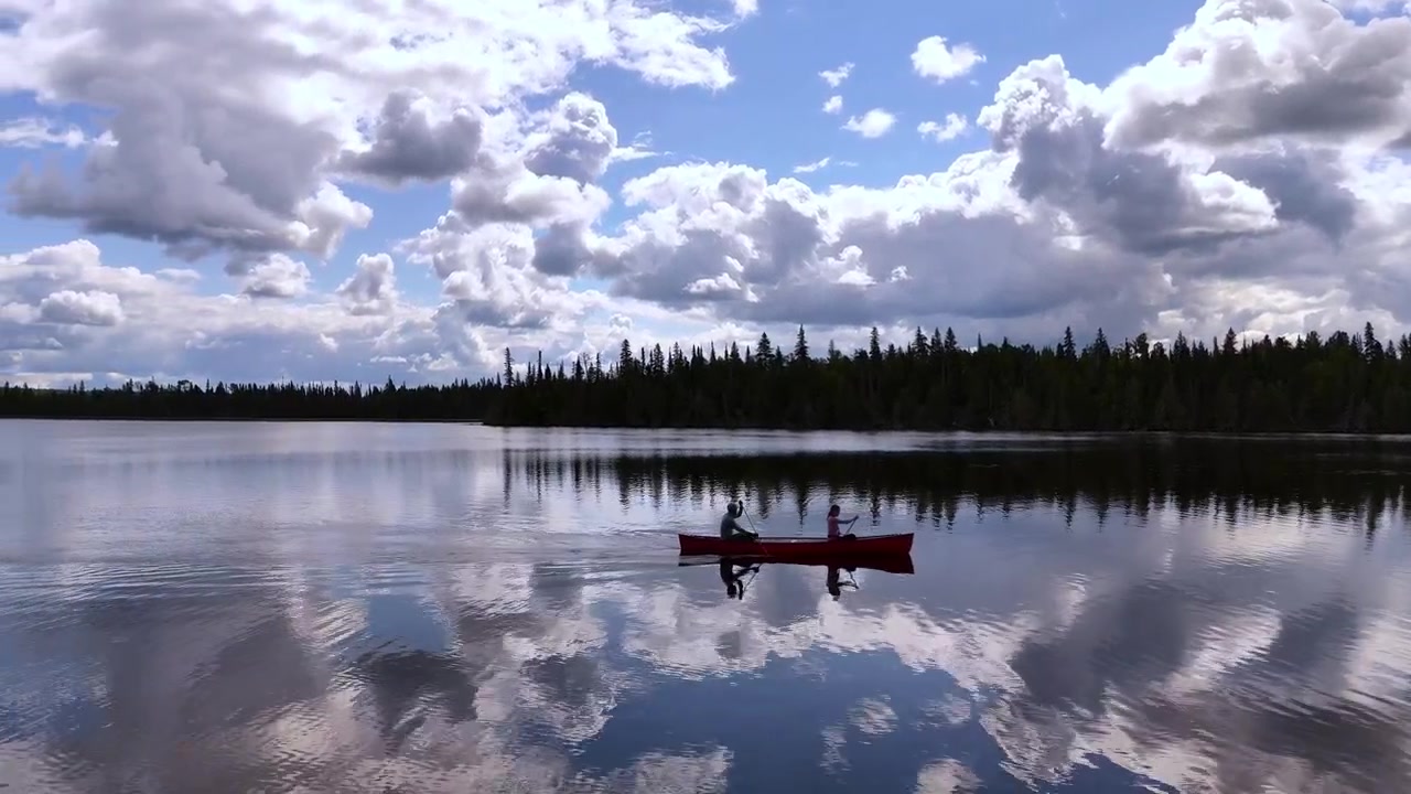 Drone follows a couple canoeing across a calm lake with mirror-like reflections of blue sky and towering clouds, creating a peaceful wilderness scene in Northern Ontario.