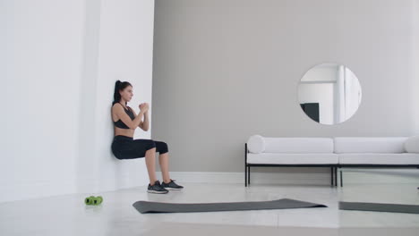 The-brunette-girl-froze-in-a-squat-leaning-on-the-wall-doing-a-static-exercise-in-a-bright-interior.-Keep-90-degree-leg-angle