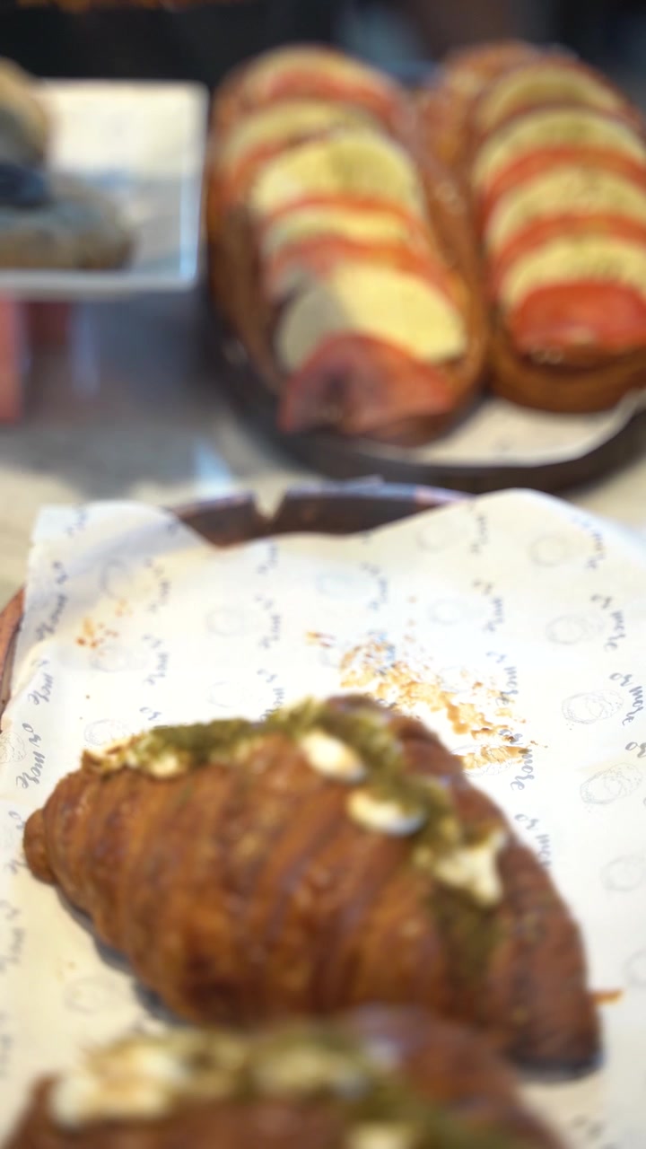Food service action. A gloved hand lifts a freshly baked, golden croissant decorated with savory toppings, with caprese sandwiches blurred in the background.