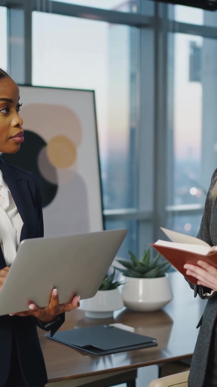Video verticale: Mostrando appunti, la donna a destra esorta una collega a controllare il laptop in ufficio per la revisione dei blazer.