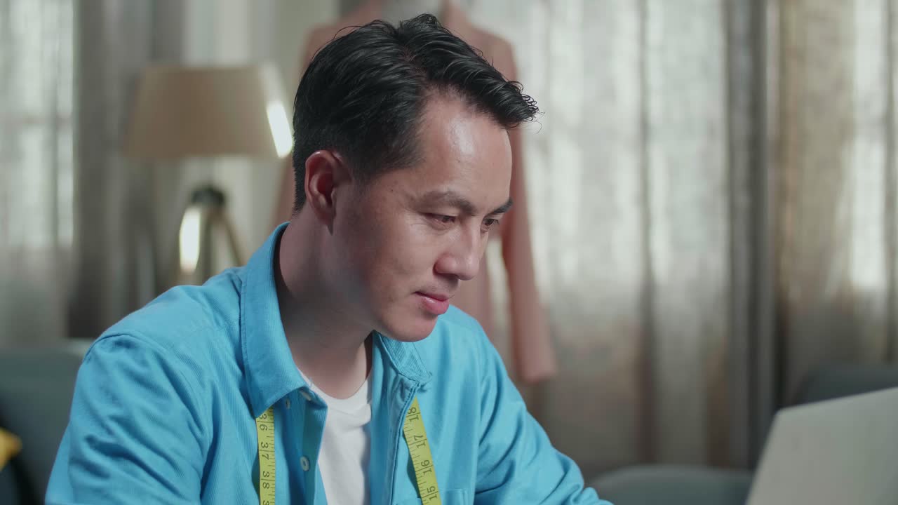 아시안 남성 디자이너가 노트북으로 작업하고 미소 짓고 스튜디오에서 옷을 디자인하는 동안 카메라로 엄지손가락을 올립니다.