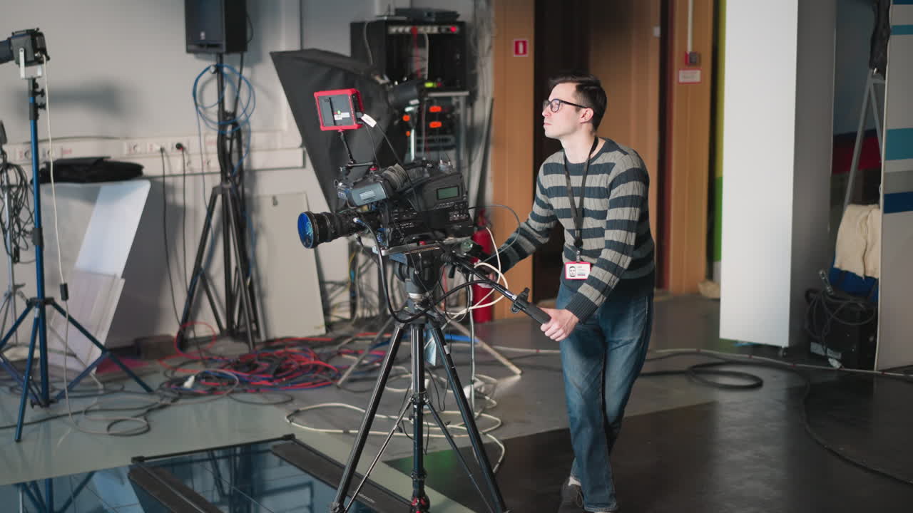Man operating professional camera in studio, adjusting camera settings while walking, surrounded by equipment, focusing on filming process, preparing for video production with a clear background