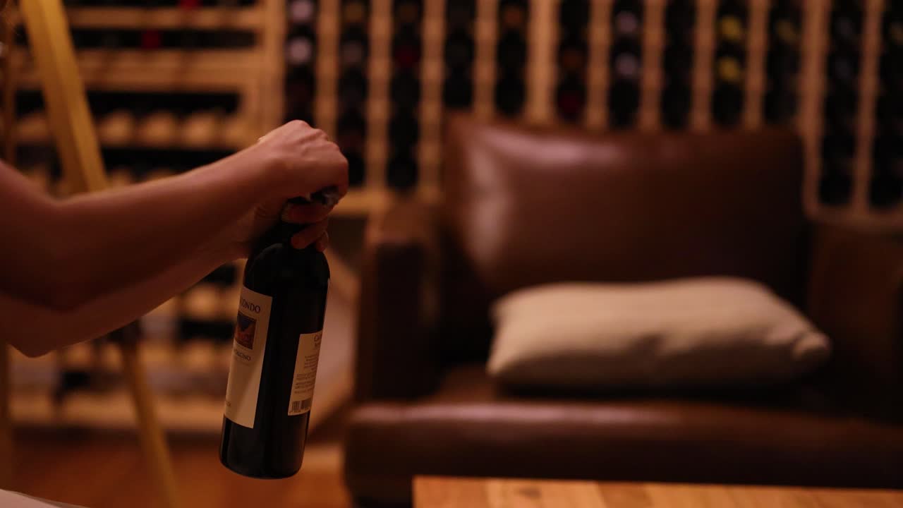 A person opens a wine bottle in a warmly lit cellar, surrounded by shelves of wine bottles and a comfortable chair