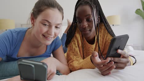 felices amigas adolescentes diversas acostadas en la cama usando teléfonos inteligentes y hablando